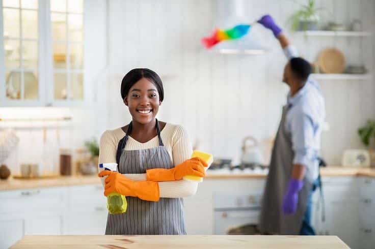 Portrait of black lady in apron and rubber gloves holding detergent, smiling at